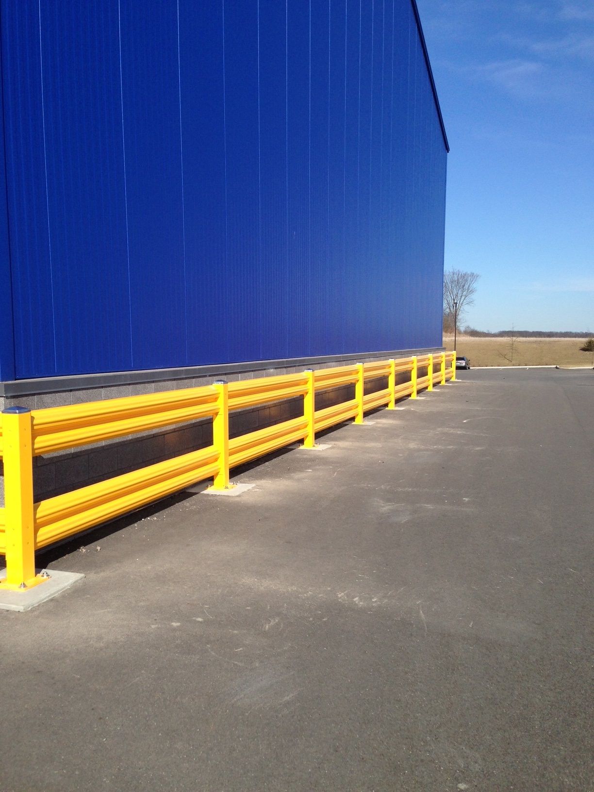 Yellow safety barriers along a blue warehouse wall on an asphalt surface. Bright, sunny day.