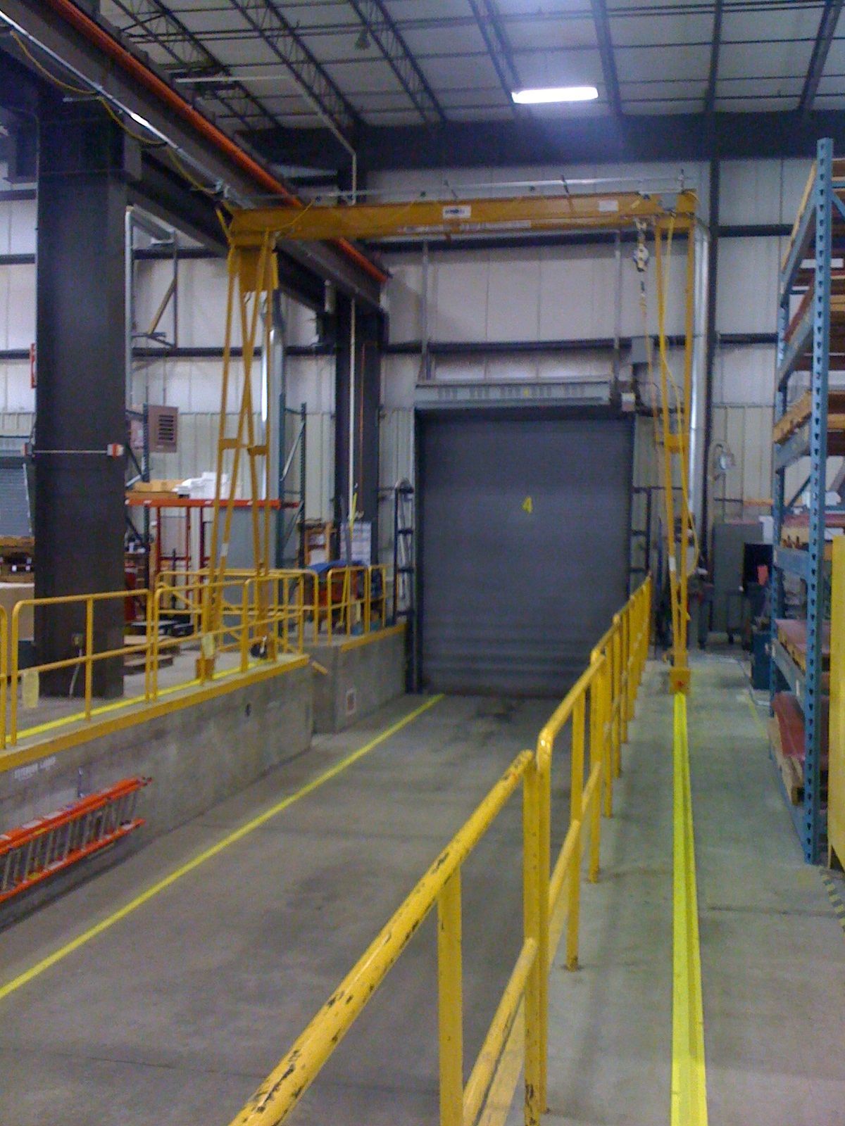 Yellow safety railings lead to a loading dock and closed gray door inside a warehouse. A yellow crane extends over the door.