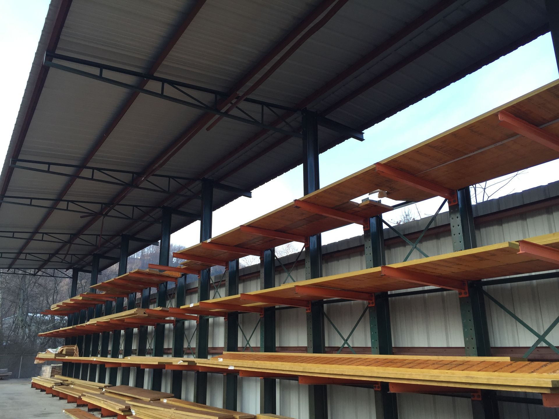 Lumber storage shed with multiple shelves holding stacks of wood. Green metal supports, gray roof, and blue sky.