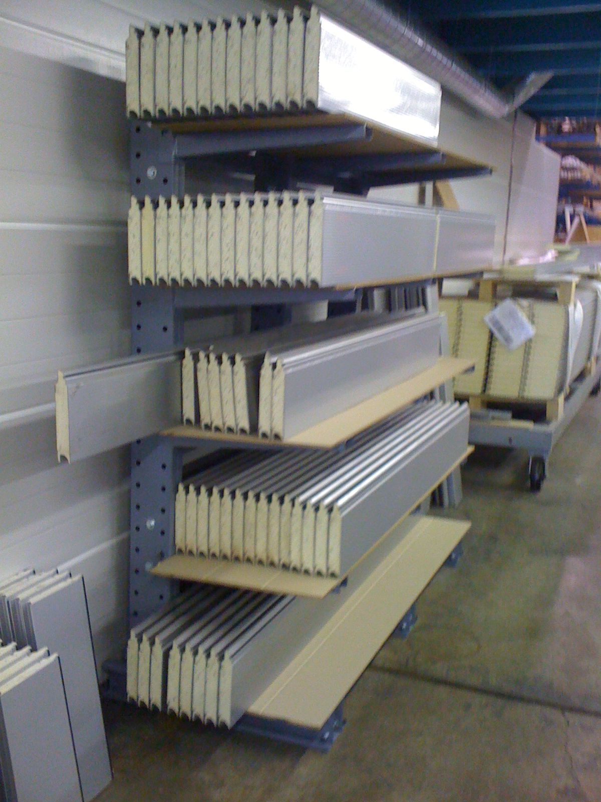 Shelving unit holding stacks of rectangular, insulated panels in a warehouse setting. The panels have a silver exterior and a white core.