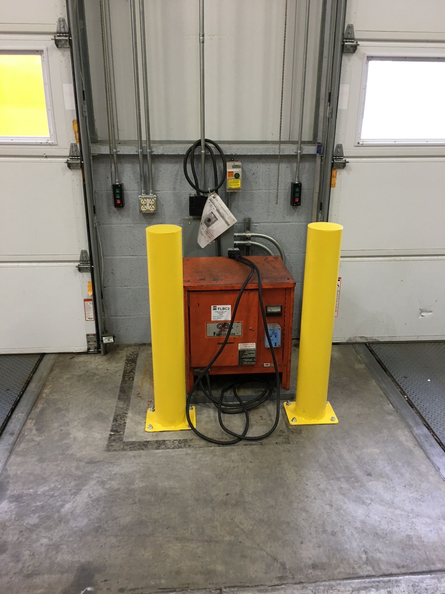 Yellow bollards protect an electrical box at a loading dock. The box is in front of a closed bay door.