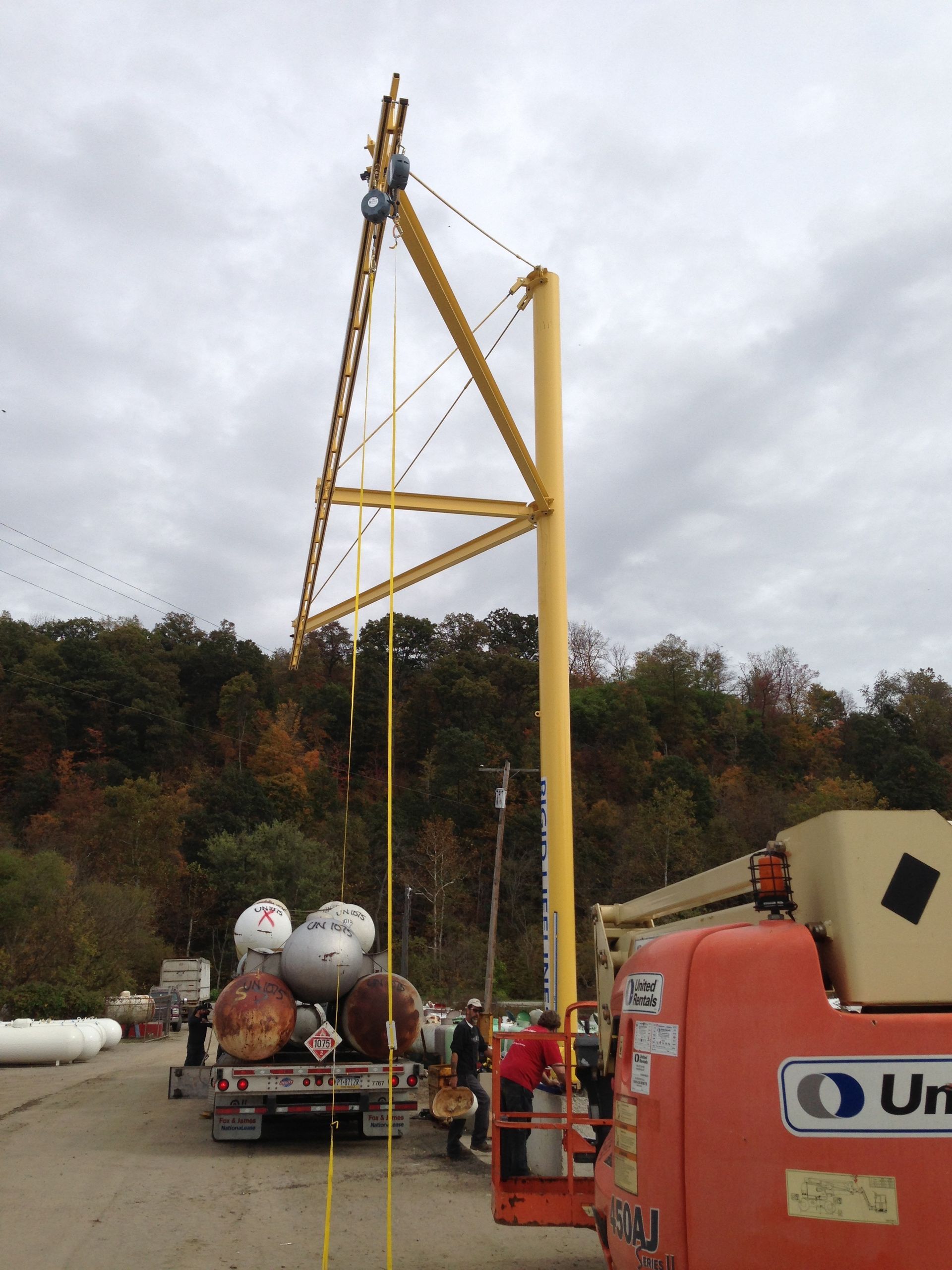 A yellow crane is lifting a tank from a truck. A worker stands in an orange lift. The setting is outdoors on a cloudy day.