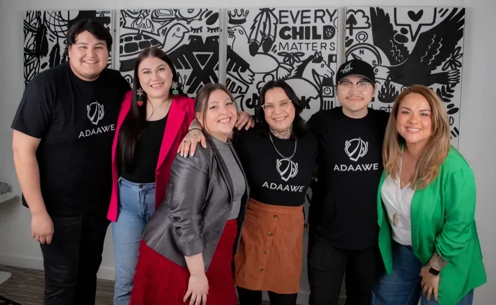 Group of six people smiling, posing in front of a black and white mural. They wear black ADAWE shirts or hats.