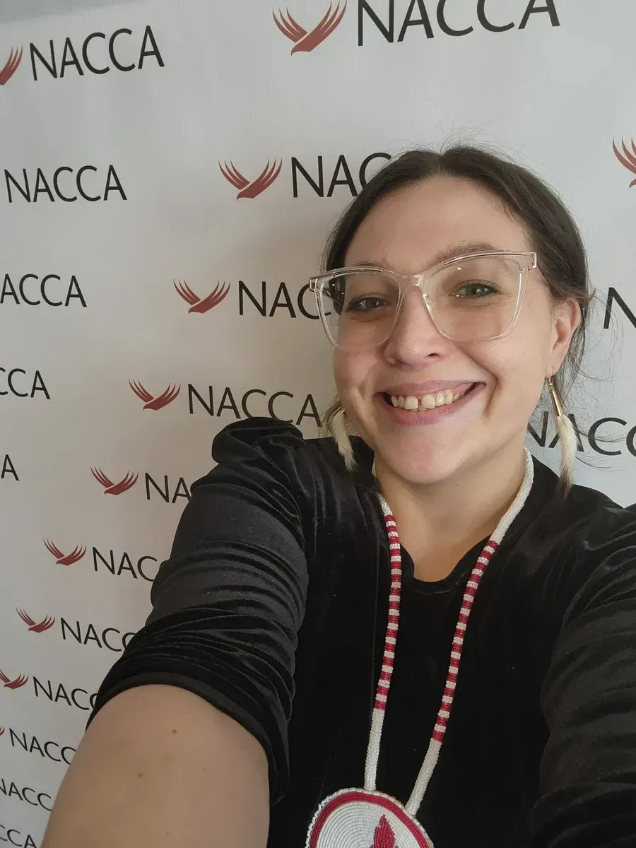 A woman wearing glasses and a beaded necklace smiles in front of a backdrop with the word 