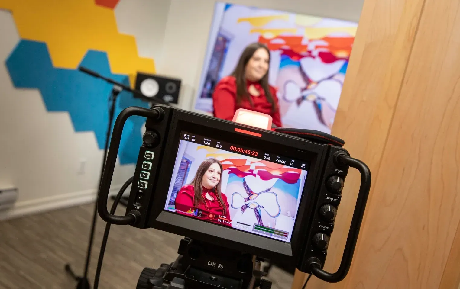 A person being filmed in a studio with colorful wall art and camera equipment. The person is wearing red.