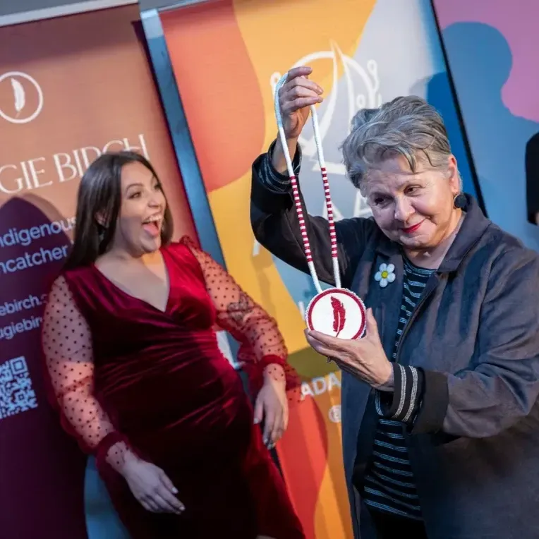 Woman holds medallion with feather design; another woman reacts excitedly.