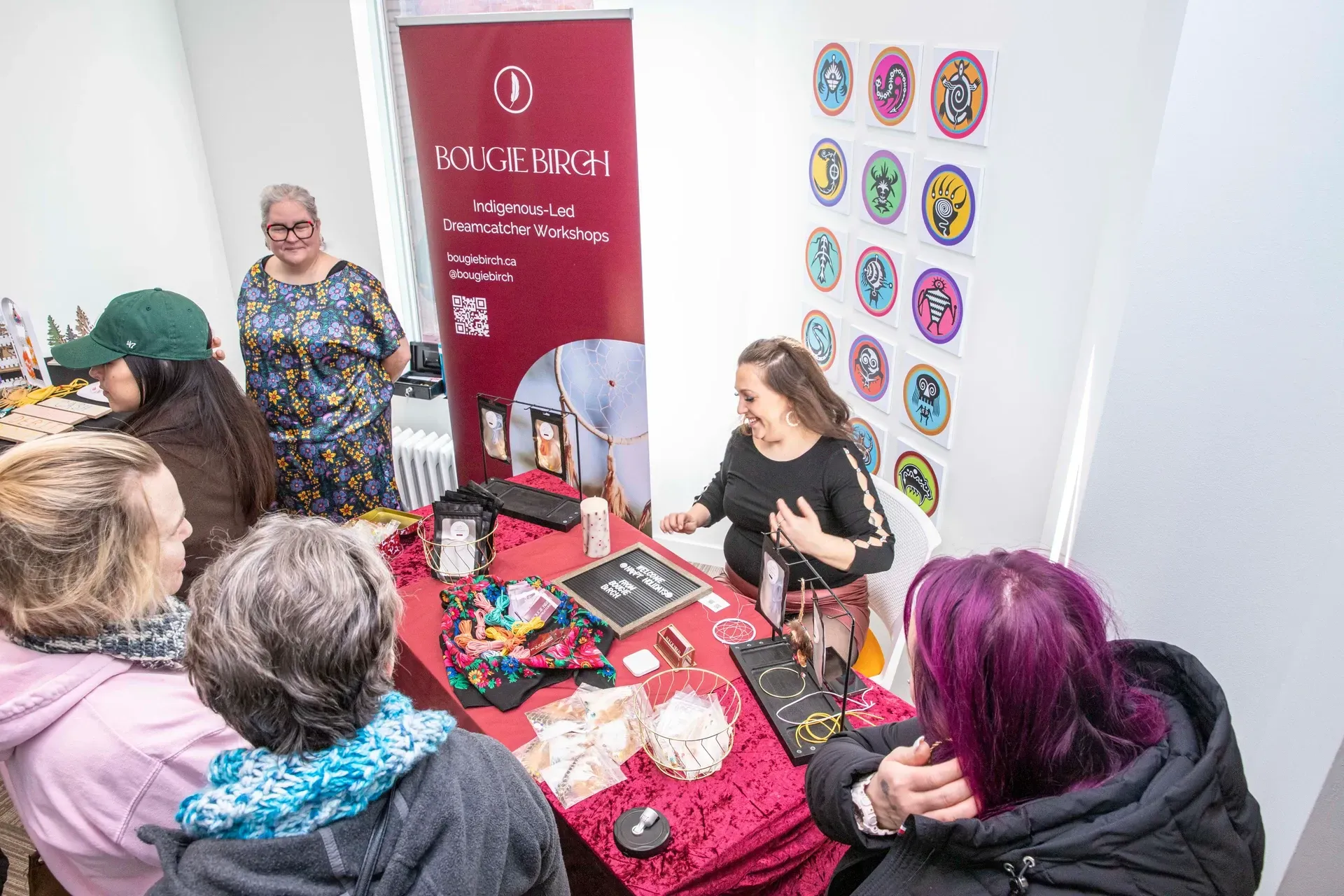 People browsing jewelry at a vendor's table inside; vendor in black top, red tablecloth, banner.