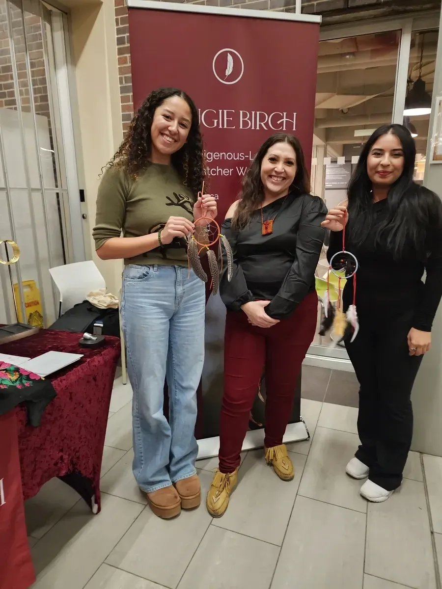 Three women stand by a display banner; one holds items. Banner says 