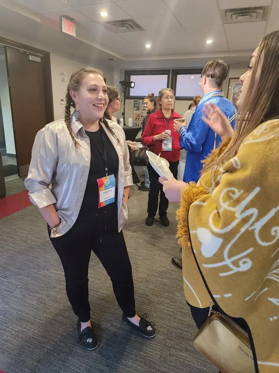 Woman in silver jacket smiles, talking with another woman. Others converse in a room.