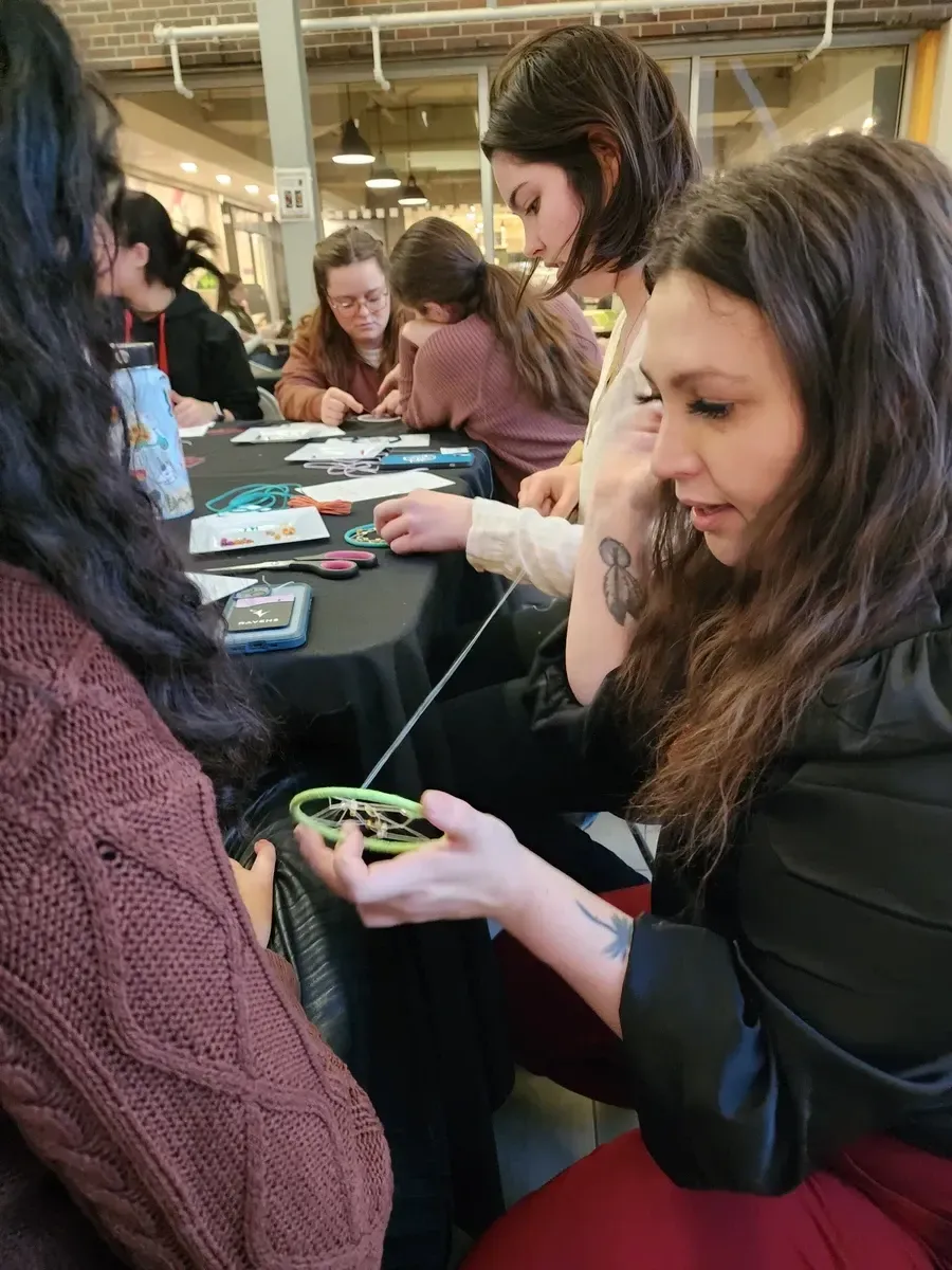 People at a table looking at cards. One woman holds a bracelet and string. Indoors with soft lighting.