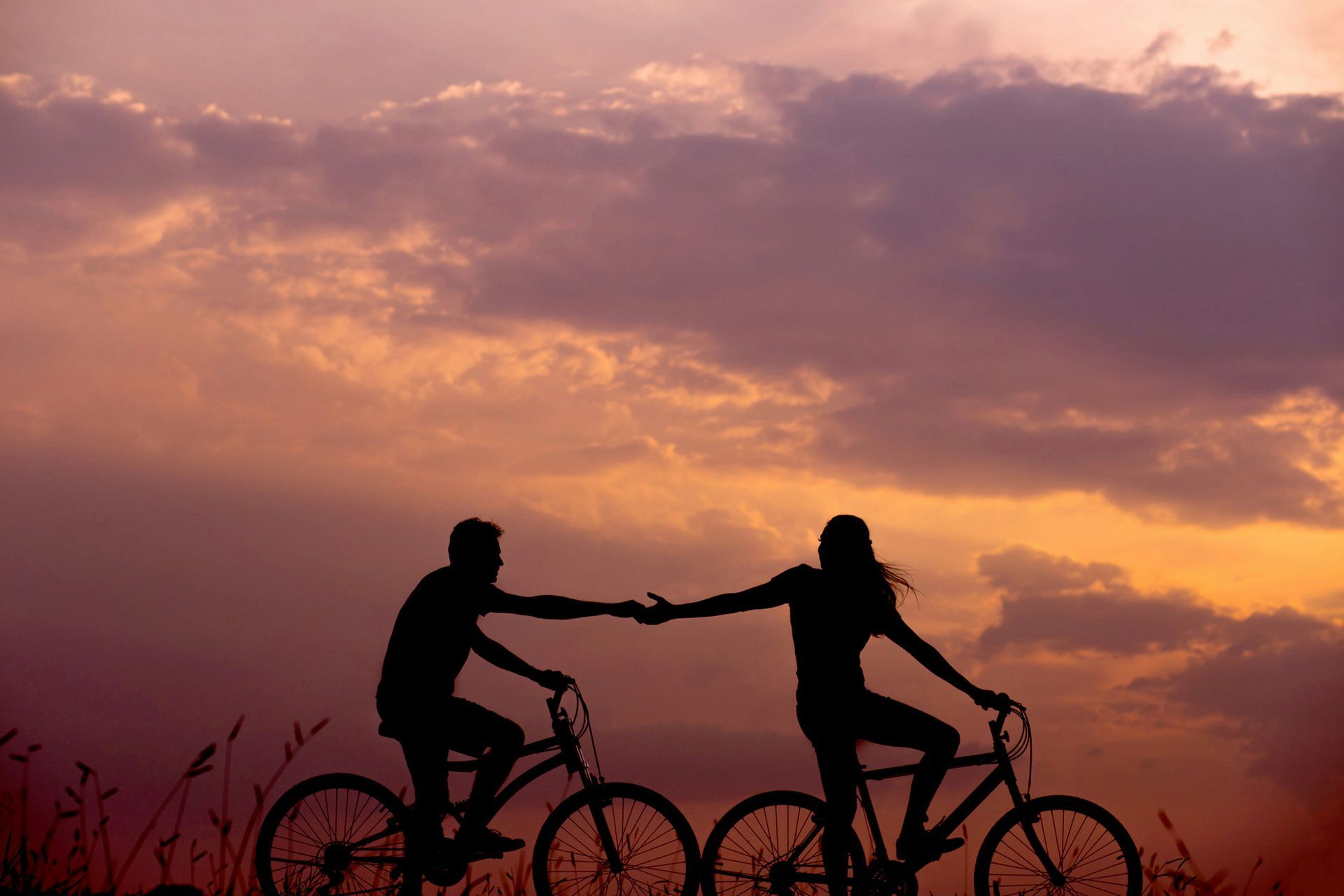 A man and a woman are holding hands while riding bicycles at sunset.