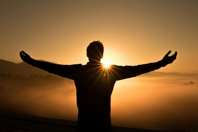 A man is standing with his arms outstretched in front of a sunset.