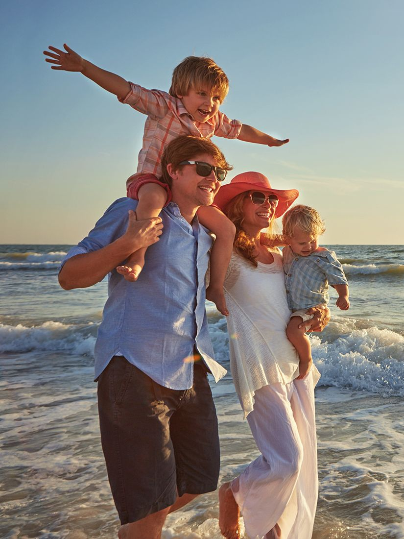 Family of four at the beach: Father carries a child, mother holds a baby; smiles, sun.