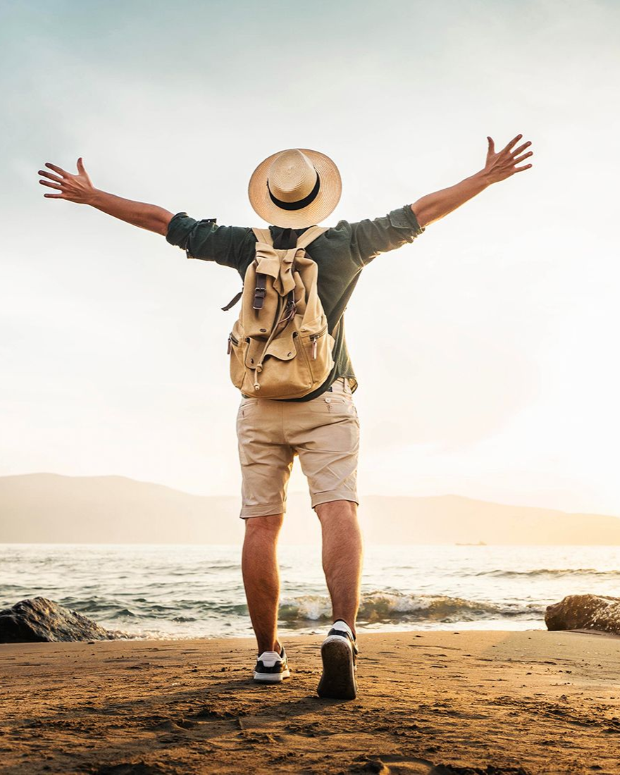 Person with arms outstretched on a beach, facing the ocean. Wearing a hat, backpack, shorts. Sunset in the background.