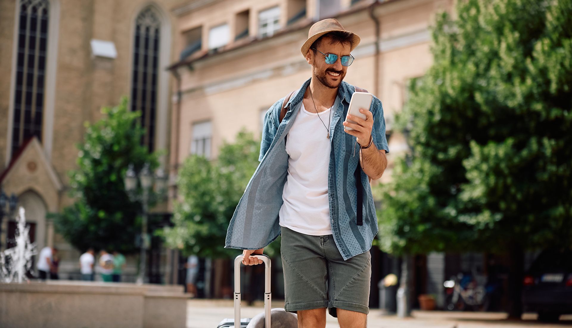 Man with suitcase, using phone, walking on a city street, wearing sunglasses, hat, shorts, and unbuttoned denim shirt.