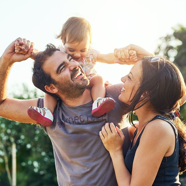 Family of three smiling outdoors; father holding child on shoulders, mother beside.