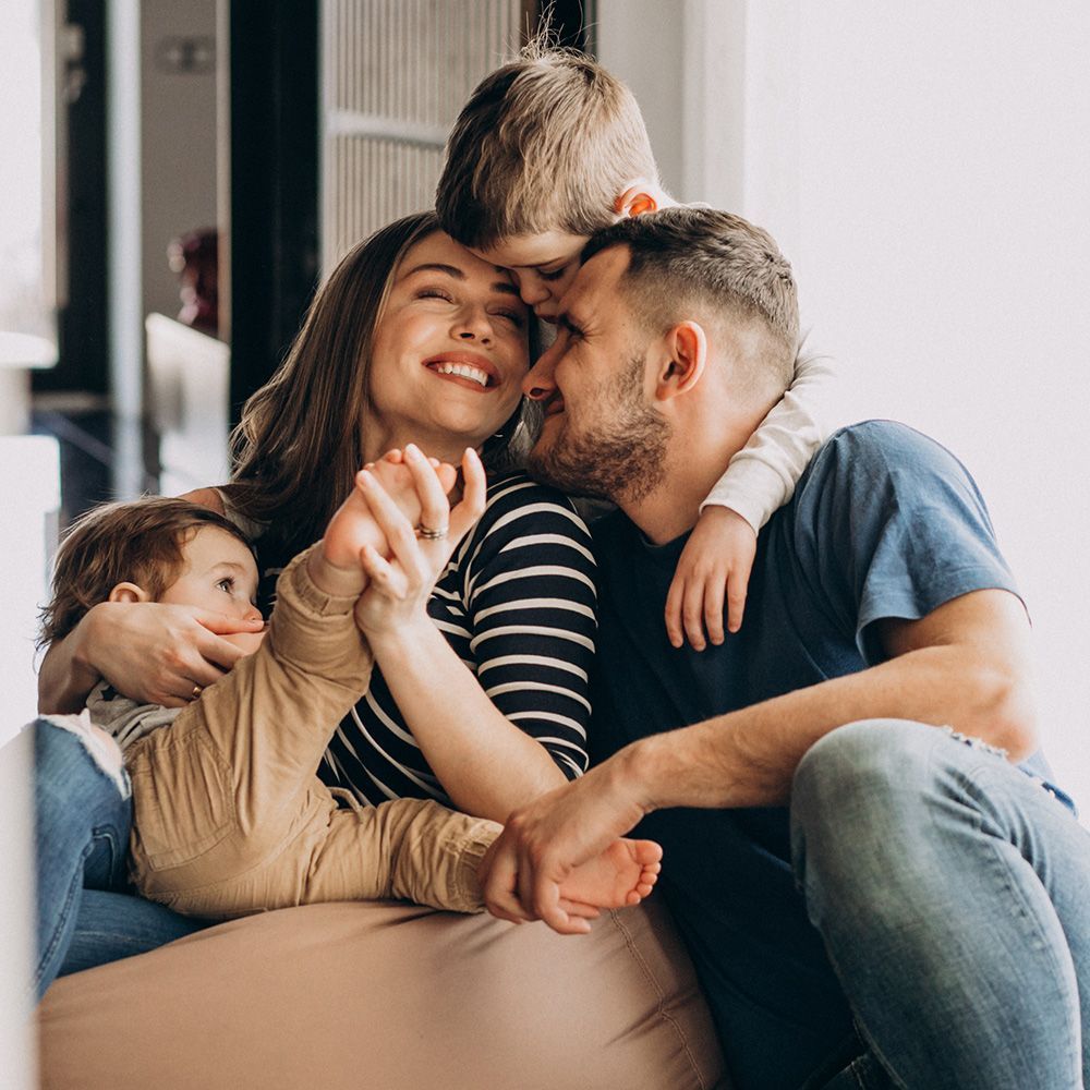 Family of four smiling and embracing, sitting by a window.