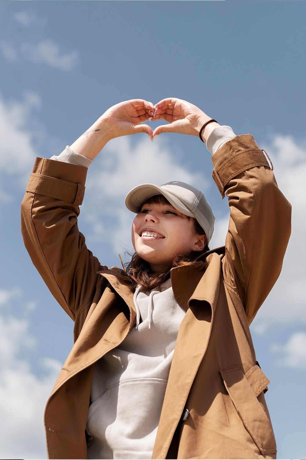 Woman forming a heart with hands against a blue sky, smiling and wearing a tan coat and hat.