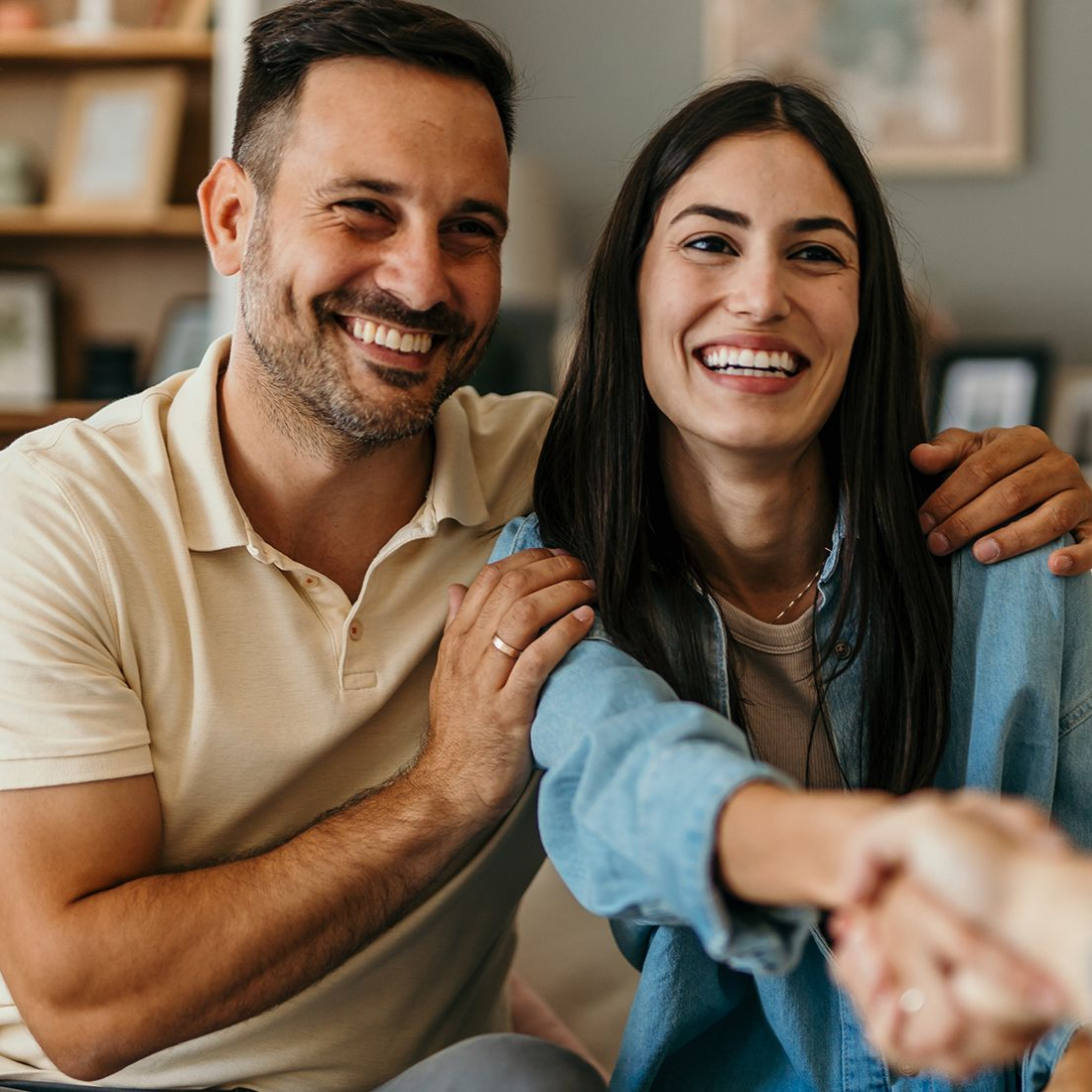 Smiling couple shaking hands with a person; indoors, man has arm around woman.