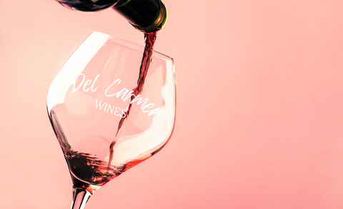 Red wine being poured from a bottle into a branded Del Carmen glass against a solid pink background.