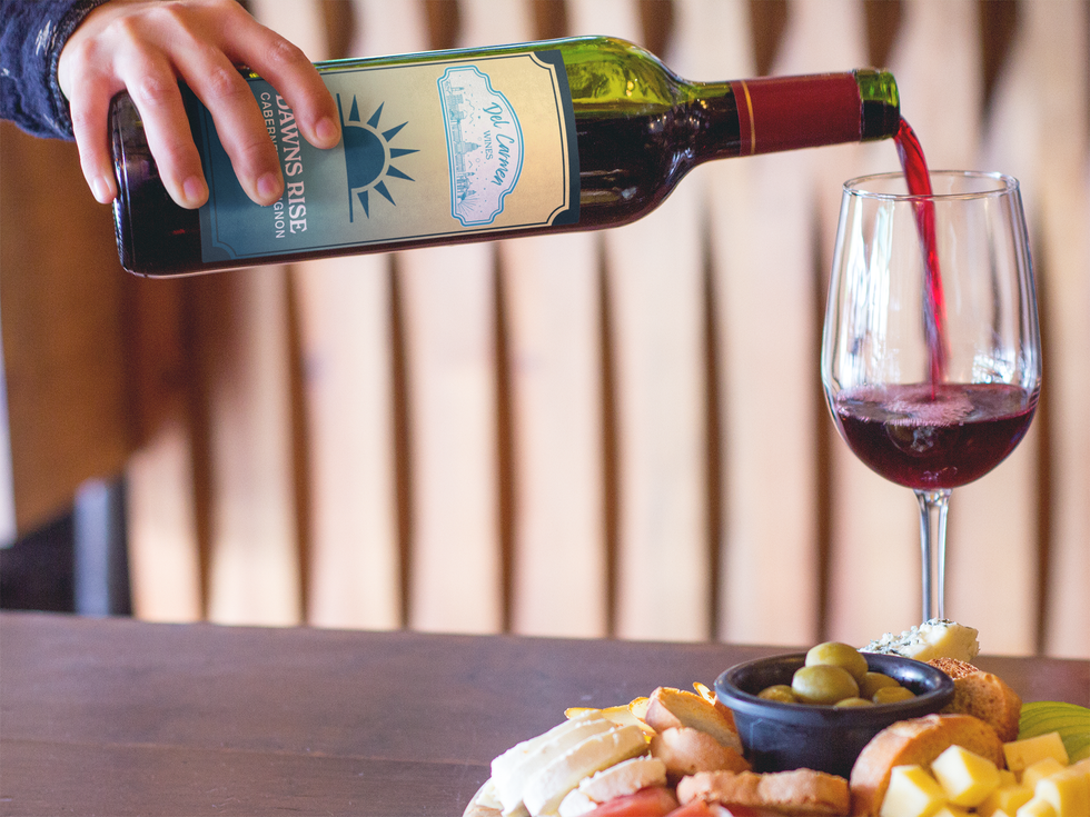 A hand pours red wine into a glass next to a small bowl of olives and a platter of cheese and crackers.