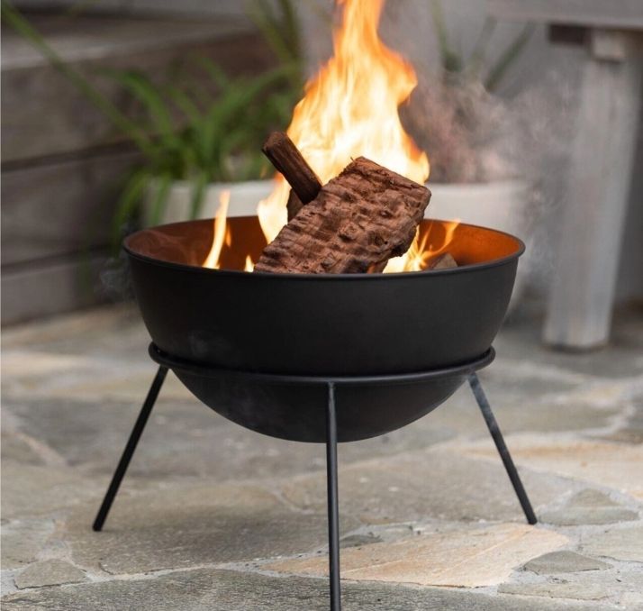 Fire bowl with burning logs on a stone patio, with a plant and chair in the background - Palms Landscape Supplies in Forster, NSW