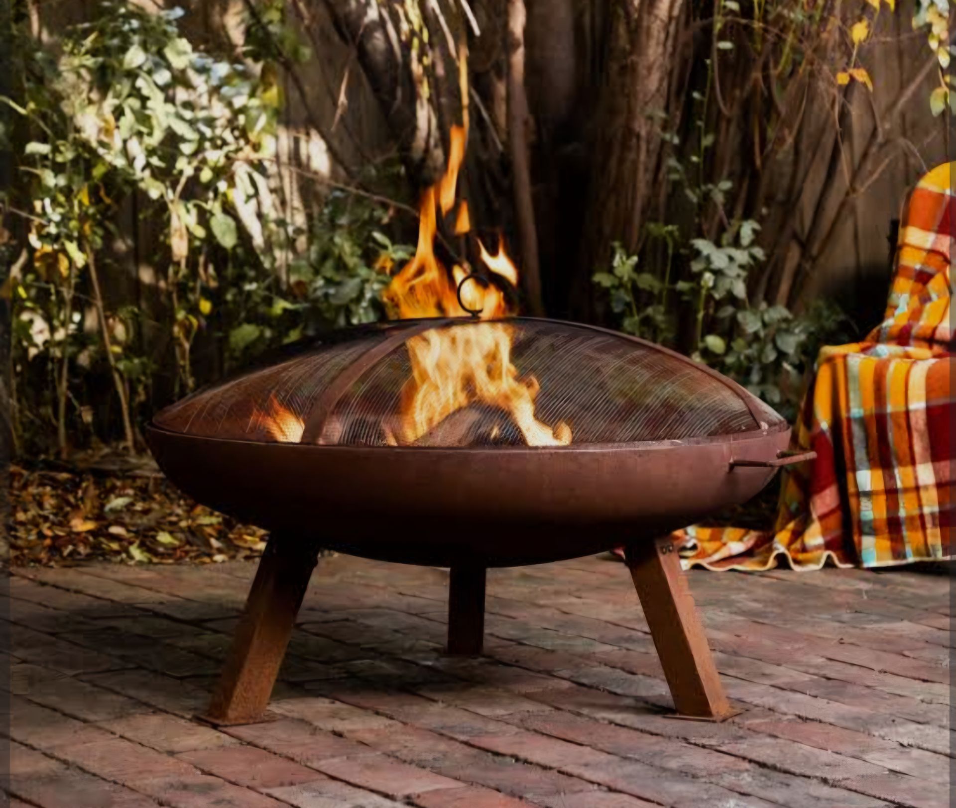 Fire pit with flames, brown metal bowl on wooden legs, screen cover, plaid blanket on chair in backyard - Palms Landscape Supplies in Forster, NSW