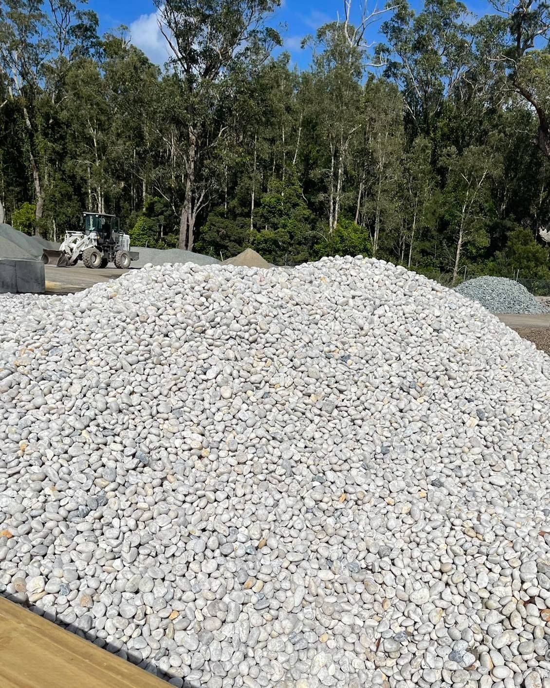Pile of light-coloured gravel in front of trees, with an excavator in the background - Palms Landscape Supplies in Forster, NSW