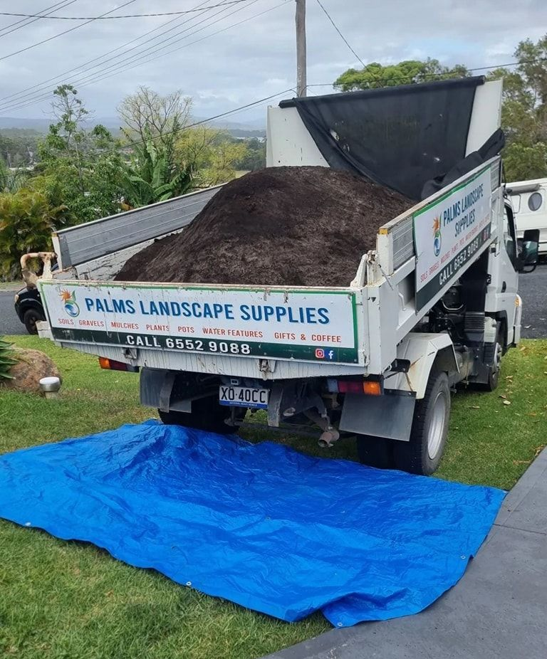 Dump truck loaded with dark soil, dumping onto a blue tarp on green grass - Palms Landscape Supplies in Forster, NSW