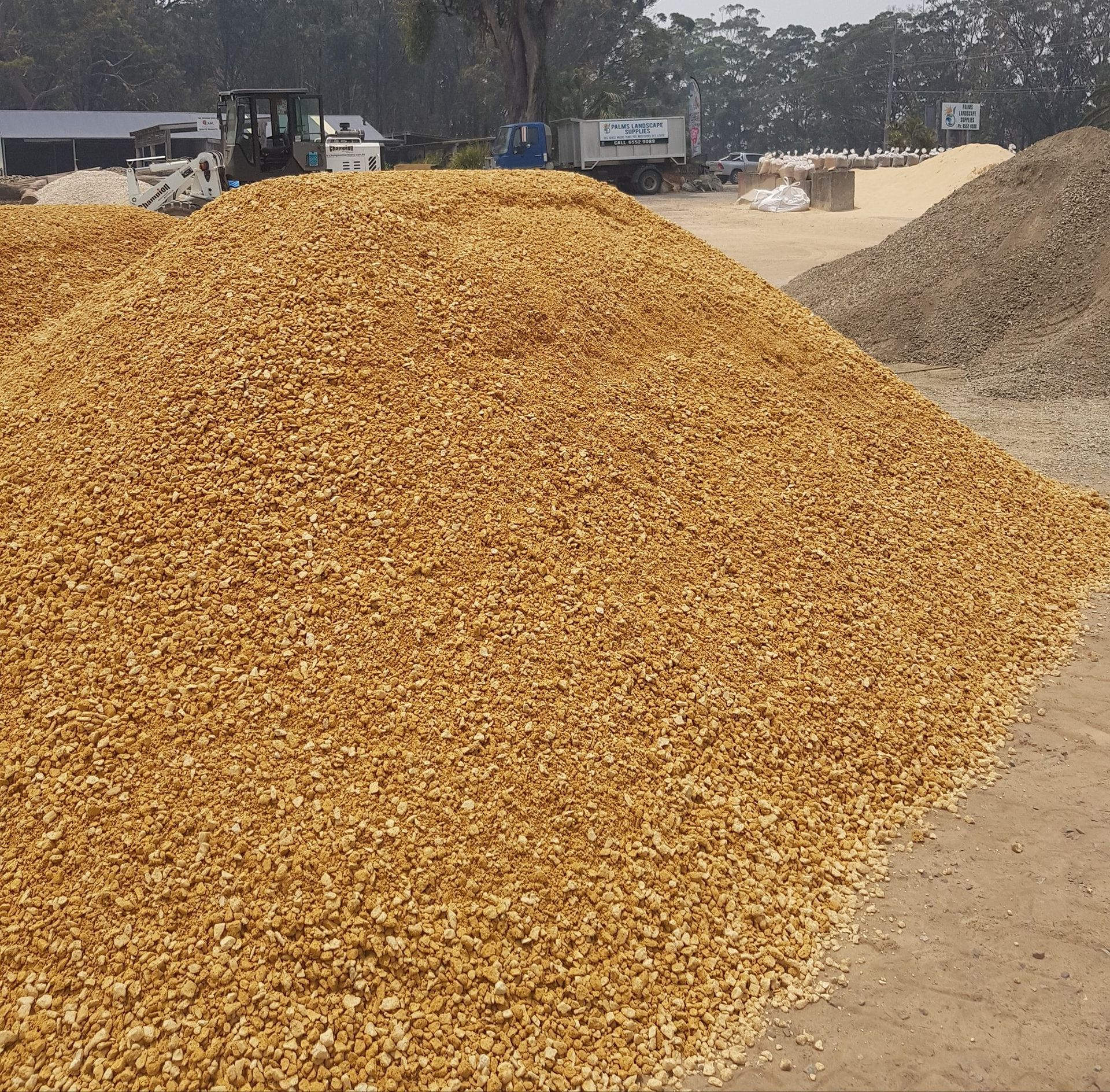 Pile of golden gravel in an outdoor setting, with a small truck and equipment visible in the background - Palms Landscape Supplies in Forster, NSW