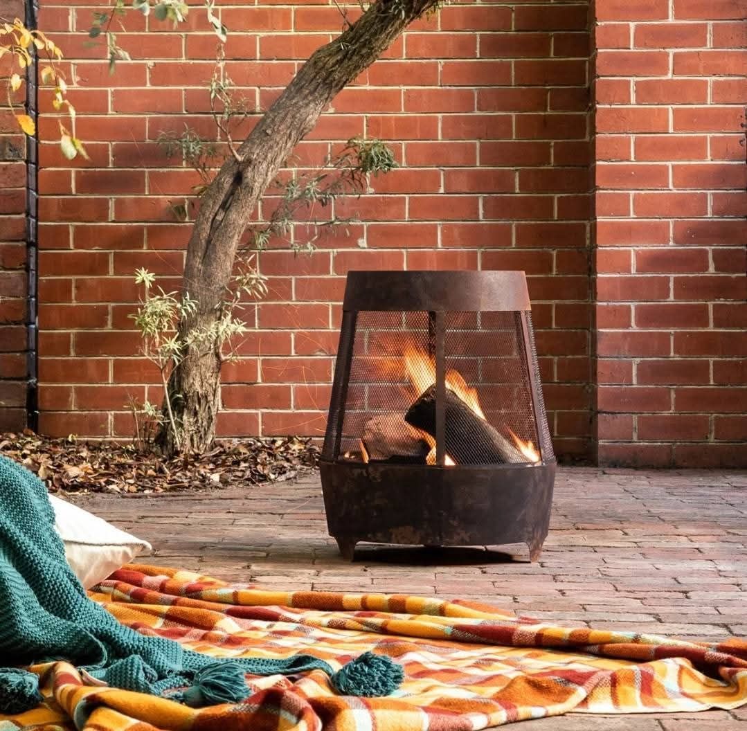 Fire pit with burning logs, on brick patio in front of a brick wall, with blanket and pillows - Palms Landscape Supplies in Forster, NSW