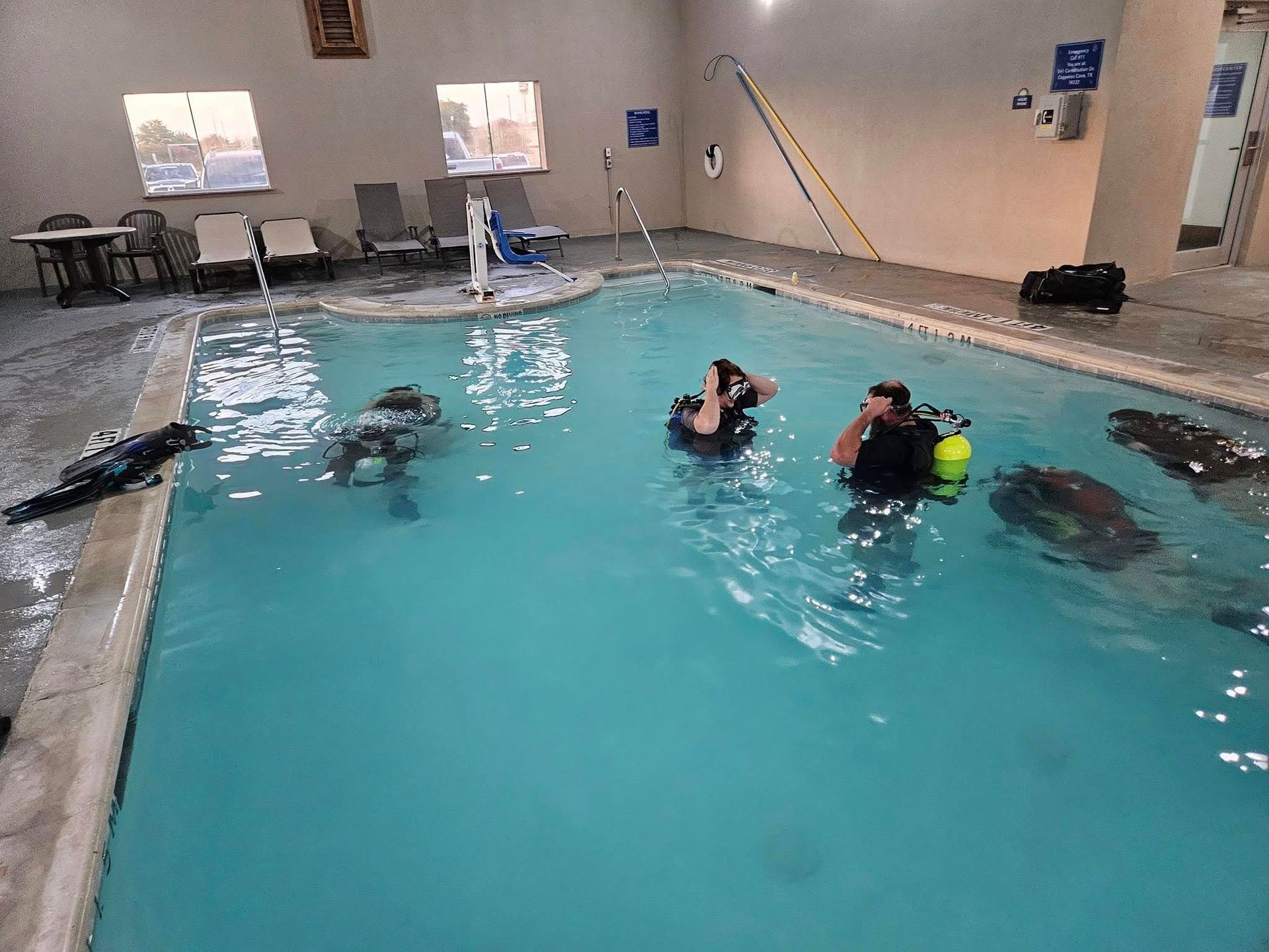 Divers in an indoor pool practice, wearing scuba gear. Others watch nearby.