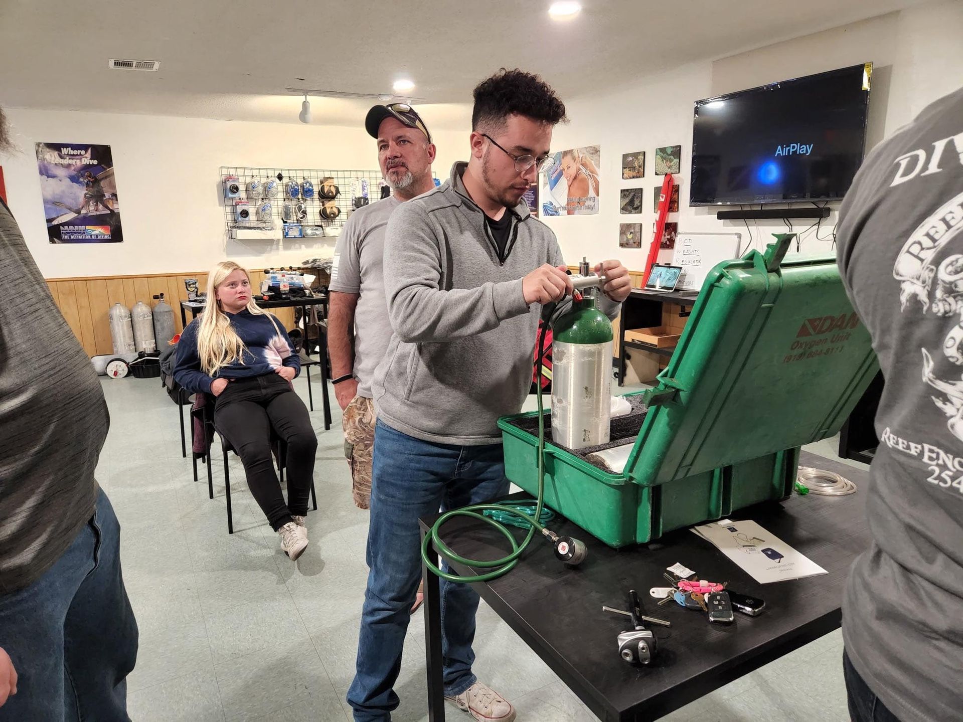 Man filling oxygen tank in a basement classroom. Others watch. Green case and tank, black table.