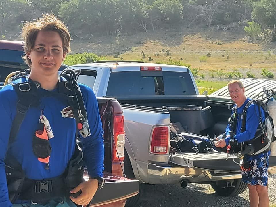 Two scuba divers by a truck, one looking at camera, preparing gear outdoors.