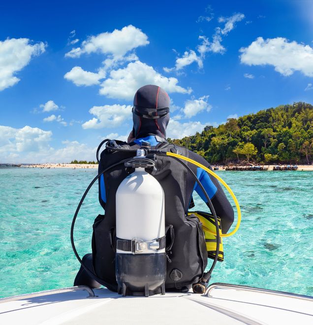 Scuba diver on boat, facing ocean, ready to dive. Clear turquoise water, blue sky, island in background.