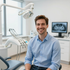 Persona sonriente en un consultorio dental con sillón, instrumental dental y monitor de rayos X al fondo.