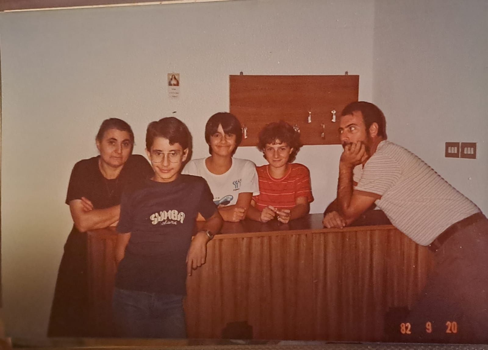Gruppo di persone a un bancone di legno. Datato 8/82. Include una donna, diversi bambini e un uomo appoggiato.
