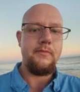 A bald man with a beard and glasses is taking a selfie on the beach.
