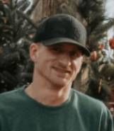 A man wearing a baseball cap and a green shirt is standing in front of a christmas tree.