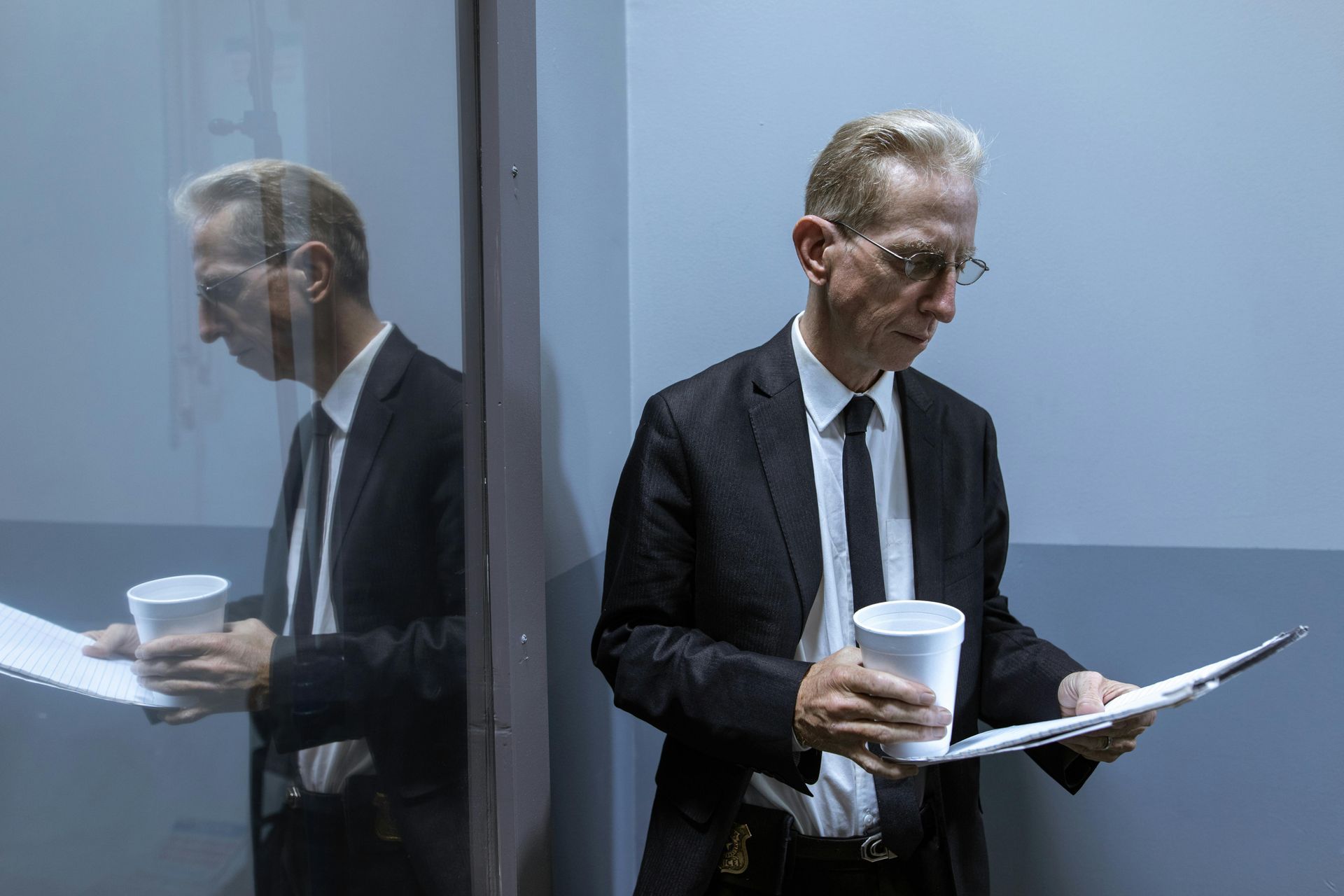 A man in a suit and tie is standing in a room holding a cup and a clipboard.