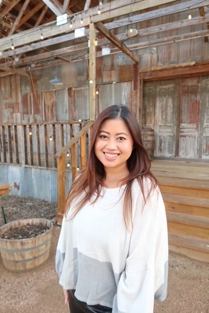A smiling person stands outside a rustic wooden barn with string lights, wearing a white and grey oversized sweater.