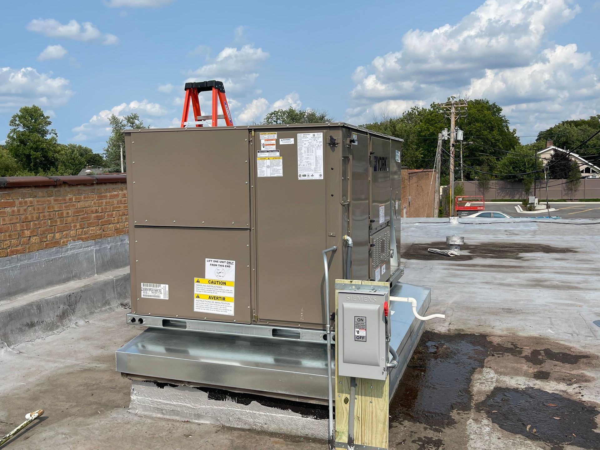 Rooftop HVAC unit. Brown and silver box on a flat roof. Ladder, electrical box, and a blue sky.
