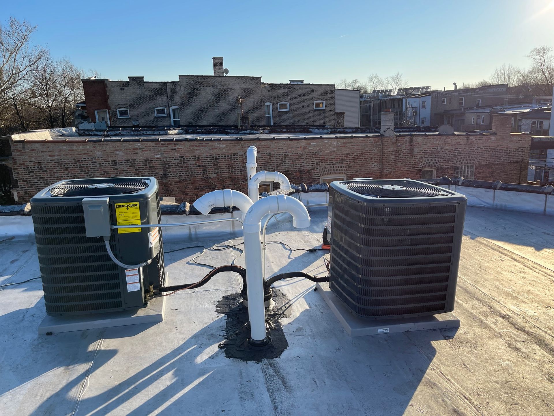 Two air conditioning units on a flat rooftop, with white pipes and a brick building in the background under a clear sky.