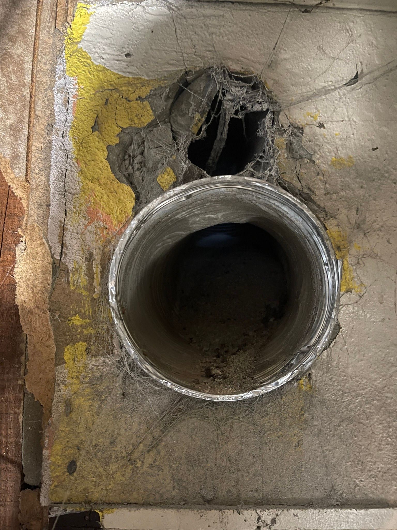 Open metal ductwork in concrete with a damaged hole above it; yellow paint is visible.