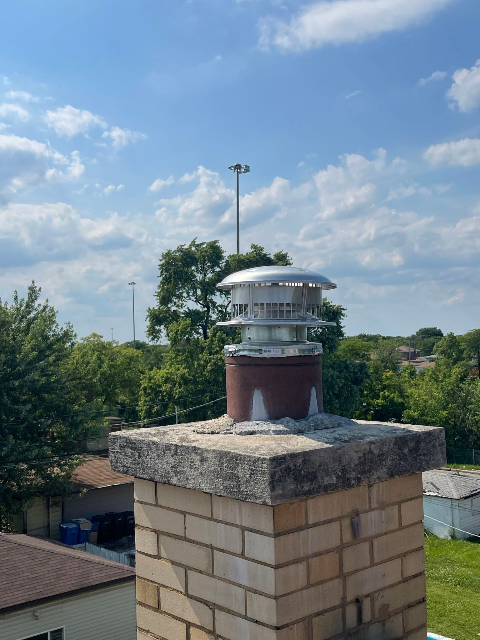Chimney with cap against a blue sky with trees and buildings in the background.