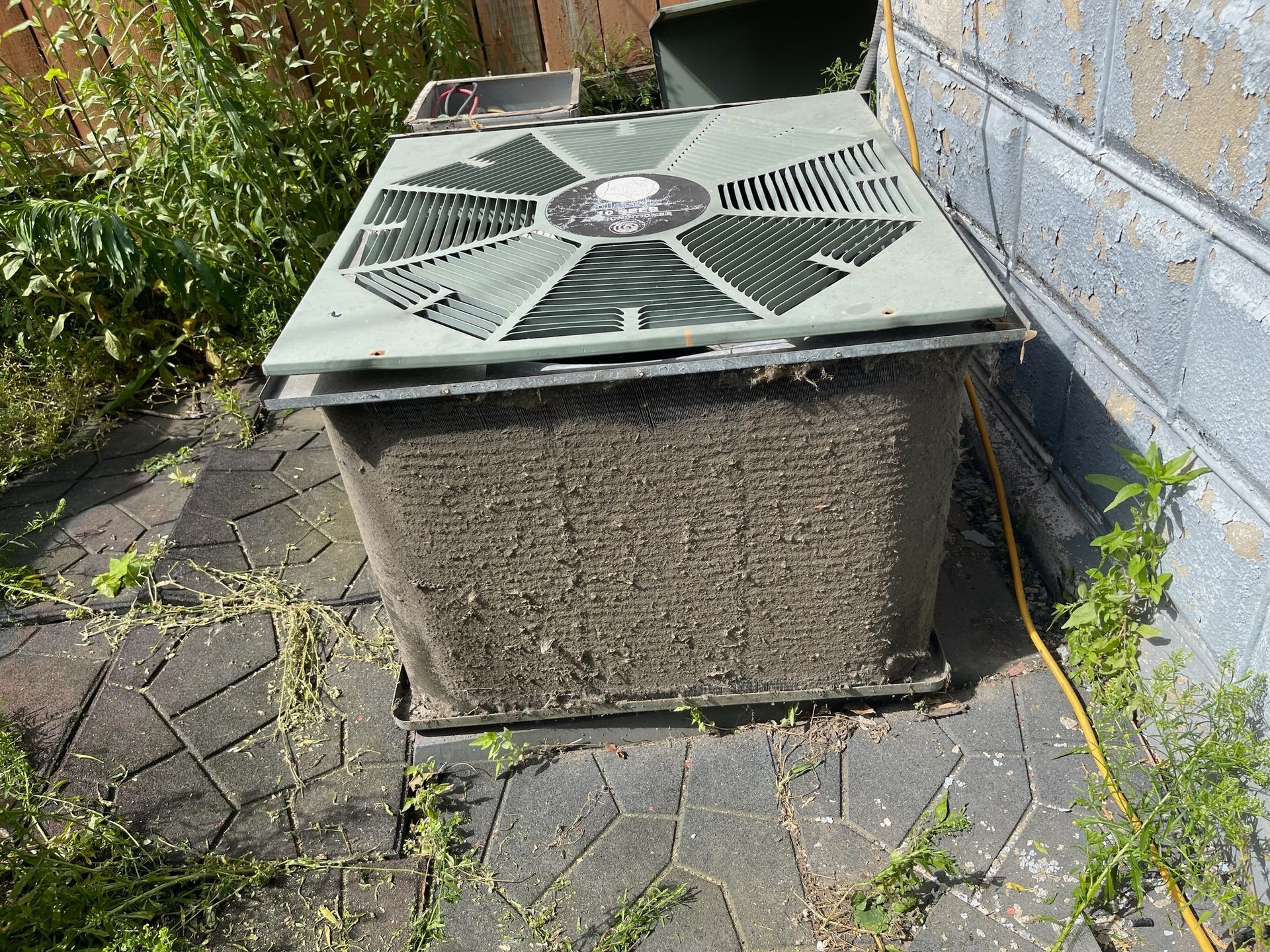 A dirty outdoor air conditioning unit sits on brick next to a wall, surrounded by some weeds.