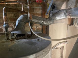 Old water heater with rusted vent pipe and copper pipes against a brick wall.