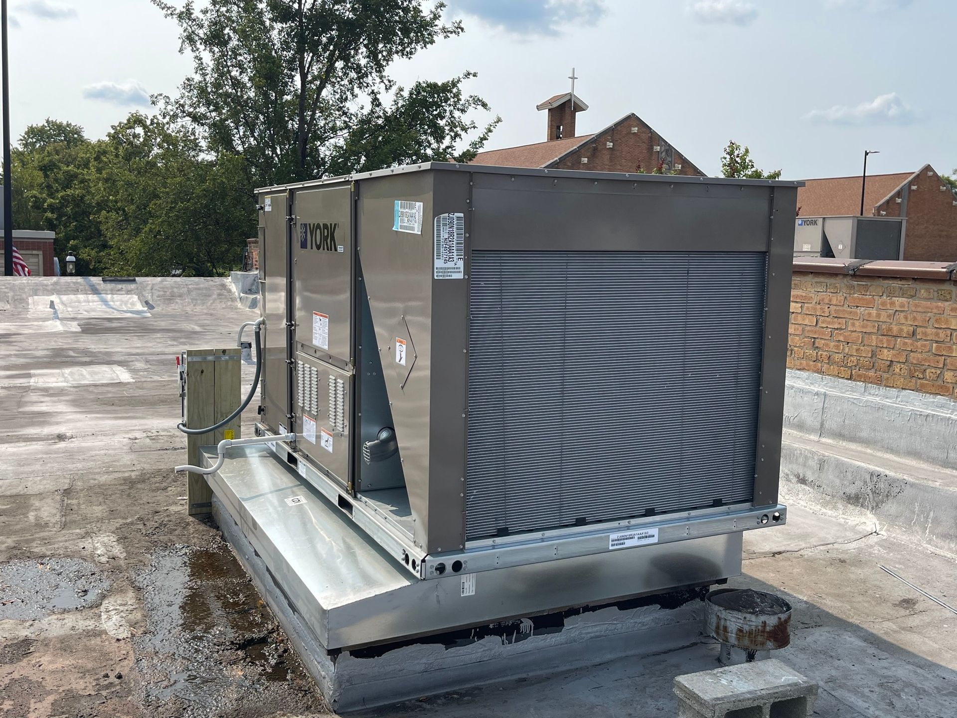 Rooftop HVAC unit, gray, on a flat roof. Brick building and trees in the background.