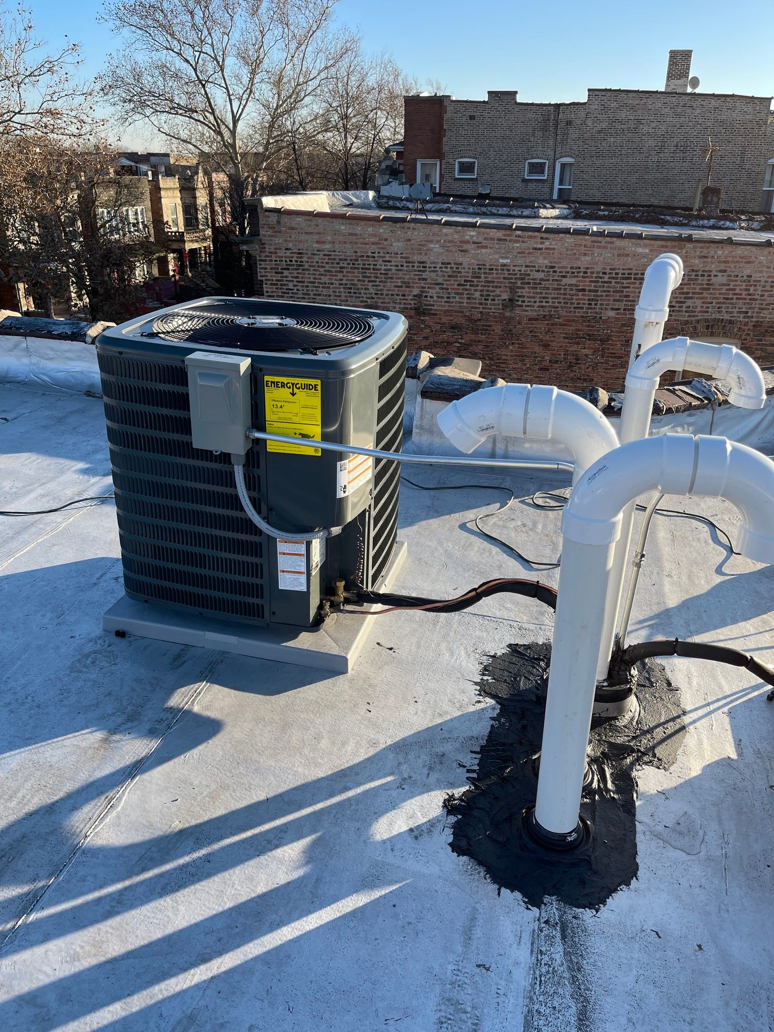 Air conditioner on a rooftop, with white pipes and city buildings in the background.