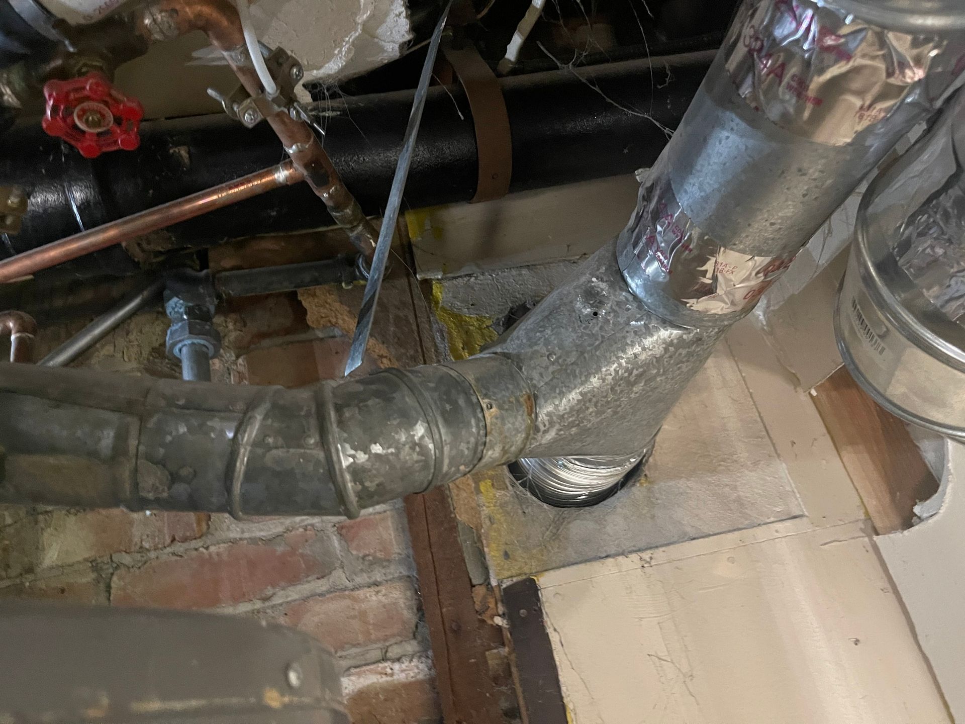Metal ductwork connection near brick wall and pipes.